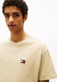 Šviesiai rudos medvilninės marškinėliai su mažu, daugiafunkciniu stačiakampio formos logotipu ant kairės krūtinės, ant kurio yra užrašas "TOMMY JEANS". Trumpomis rankovėmis, apvaliu kaklu.