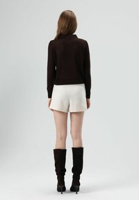 Bruine gebreide trui met kraag, gecombineerd met witte korte broek en kniehoge donkerbruine laarzen. De outfit heeft een aansluitende pasvorm.