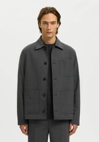 Tumši pelēks žakets ar apkakli, pogu aizdari un divām lielām priekšējām kabatām. Gluda tekstūra ar minimālām šuvju detaļām.