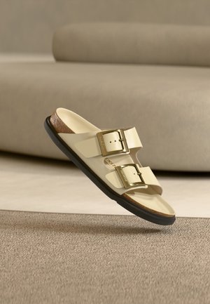 Helle beiger Sandale mit zwei verstellbaren Riemen und goldenen Schnallen. Strukturierte schwarze Sohle und brauner Akzent an der Ferse. Glatte Oberflächengestaltung.