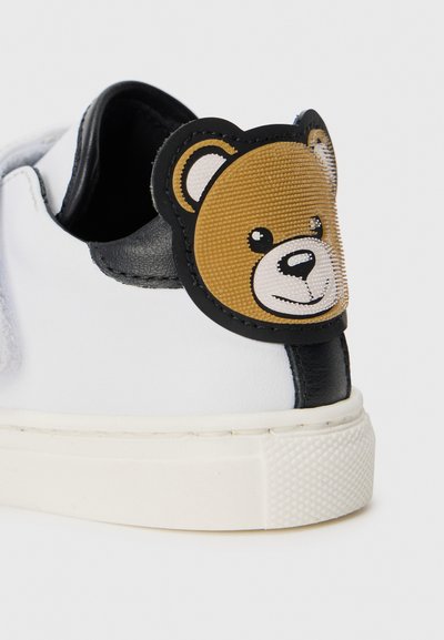 White sneaker with black leather accents and a textured bear face patch on the heel. Rubber sole with a patterned grip.