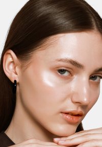 Boucles d'oreilles cerclées noires et brillantes portées par un modèle à la peau lisse et au maquillage léger, avec une texture douce et naturelle et des cheveux bruns raides.