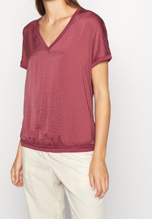 Mujer con una camiseta de manga corta texturizada color granate con escote en V y pantalones beige claros frente a un fondo claro y liso.