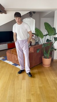 Világos lila, gumis derekú nadrág fehér pólóval párosítva, fekete cipővel viselve. A szobában barna kanapé és zöld növény található.