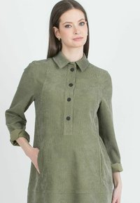 Abito in corduroy verde oliva con collo, chiusura a bottoni frontale e tasche laterali. Tessuto strutturato con polsini arrotolati.