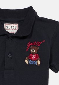 Μπλε μπλουζάκι πόλο με ριμπ γιακά, με κεντημένη αρκούδα που φοράει κόκκινο πουλόβερ και μπλε τζιν, και το λογότυπο «guess» με κόκκινα γράμματα.