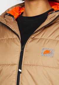 Brun puffjacka med en orange innerfoder, dragkedja fram, och en genomskinlig Nike-logotyppatch med prickar på bröstområdet.