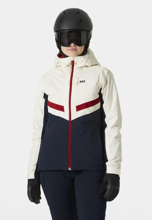 Ski jacket in cream, navy, and red with a chevron design. Features a hood, front zip, and black gloves. Worn with a helmet and goggles.
