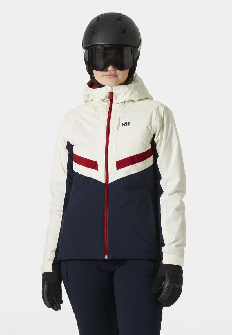 Ski jacket in cream, navy, and red with a chevron design. Features a hood, front zip, and black gloves. Worn with a helmet and goggles.