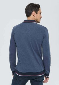 Homme portant un polo à manches longues bleu marine avec un col et des poignets rayés, de dos contre un fond clair uni.