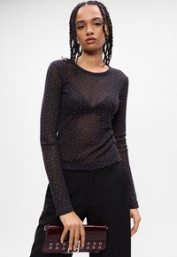 Top negro de manga larga hecho de tela transparente con puntos plateados dispersos. Escote redondeado y estilo ajustado, combinado con un bolso burdeos.