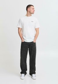 Camiseta blanca de algodón con un pequeño gráfico, combinada con pantalones acanalados oscuros de corte holgado y zapatillas blancas. Diseño simple y casual.