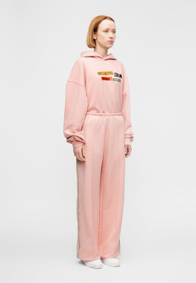 Ροζ φούτερ με κουκούλα και γραφικό κείμενο, συνδυασμένο με ταιριαστό ροζ παντελόνι φόρμας με ρίγες στο πλάι. Μαλακό ύφασμα, άνετη εφαρμογή, ελαστική μέση.