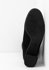 Schwarze Wildleder-Stiefelette mit einem niedrigen Absatz. Die Sohle weist geprägte Muster und Logos auf, die strukturiertes Material und Designdetails zeigen.