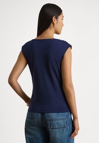 Granatowy top bez rękawów z okr�ągłym dekoltem, z teksturowanymi akcentami na ramionach i dopasowanym krojem. W połączeniu z jasnoniebieskimi dżinsami.