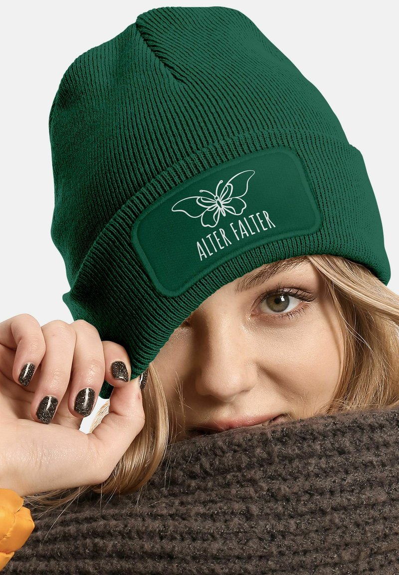 Green ribbed knit beanie with a rectangular green patch displaying a white butterfly logo and the text "ALTER FALTER." Lying against a brown scarf.