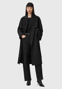 Una donna con capelli neri lisci indossa un lungo trench coat nero, una maglietta nera, jeans neri e stivaletti neri con tacco a punta, in piedi contro uno sfondo grigio.