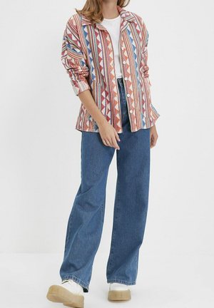 Person trägt ein mehrfarbig gestreiftes Hemd mit Knöpfen, ein weißes T-Shirt, weite blaue Jeans und weiße Plateauschuhe und steht vor einem weißen Hintergrund.