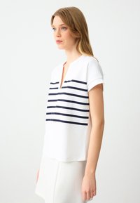 Junge Frau mit blonden Haaren, die ein weißes T-Shirt mit kurzen Ärmeln und navyblauen horizontalen Streifen sowie einem V-Ausschnitt trägt. Sie steht vor einem schlichten weißen Hintergrund.