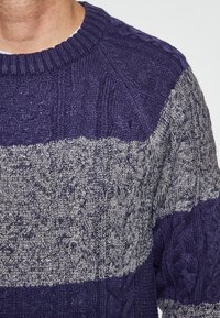 Kötött pulóver bordázott kerek nyakkal, váltakozó sötétkék és világosszürke fonott mintákkal. Texturált anyag az egész felszínen.