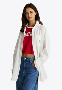 Hvid skjorte med knapper lagdelt over en rød cropped top med hvid tekst, parret med højtaljede blå denim jeans.