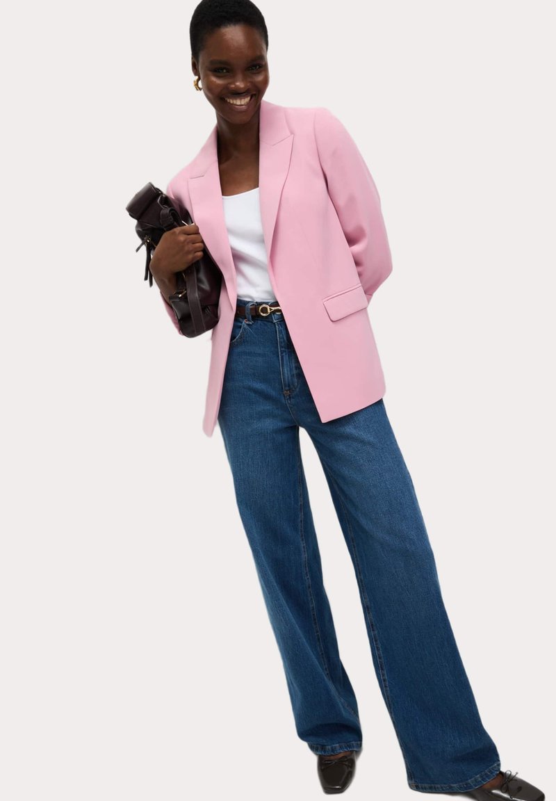Blazer ajusté rose clair sur un haut blanc, associé à un jean bleu à jambes larges et des chaussures noires. Tenant un sac à main sombre.