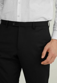 Svarta byxor i slät tyg, med knäppning, bälteshällor och skräddarsydd passform. Bärs med en vit skjorta.