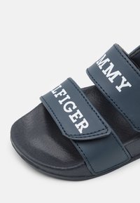 Ciemnoniebieskie klapki z dwoma regulowanymi paskami. Biały napis "TOMMY HILFIGER". Teksturowana czarna podeszwa zapewniająca przyczepność.