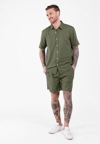 Uomo sorridente con tatuaggi su braccia e gambe, che indossa una camicia a maniche corte verde oliva con bottoni, pantaloni corti abbinati e sneakers bianche su sfondo bianco.