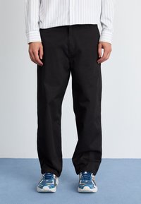 Schwarze Hose mit geradem Bein, aus einem glatten Stoff gefertigt. Kombiniert mit blauen Sportschuhen, die eine Mischung aus Texturen und Farben aufweisen.