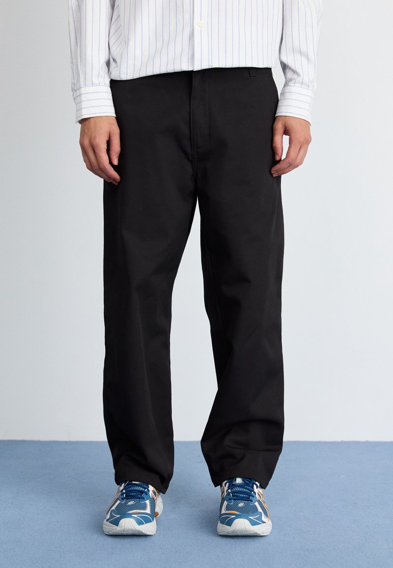 Schwarze Hose mit geradem Bein, aus einem glatten Stoff gefertigt. Kombiniert mit blauen Sportschuhen, die eine Mischung aus Texturen und Farben aufweisen.