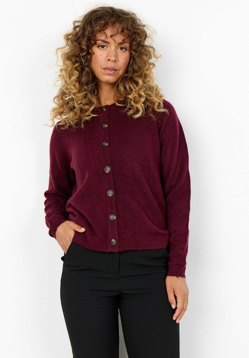 Donna dai capelli ricci che indossa una cardigan bordeaux con bottoni e pantaloni neri, in piedi con una mano in tasca su uno sfondo bianco.
