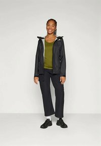 Smilende kvinne som står iført en svart hettejakke over en olivengrønn skjorte, sorte bukser, hvite sokker og svarte joggesko.