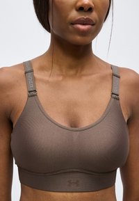 Γυναίκα που φοράει καφέ αθλητικό σουτιέν Under Armour με ρυθμιζόμενους ιμάντες, φωτογραφημένη από τους ώμους μέχρι την κοιλιά σε απλό φόντο.