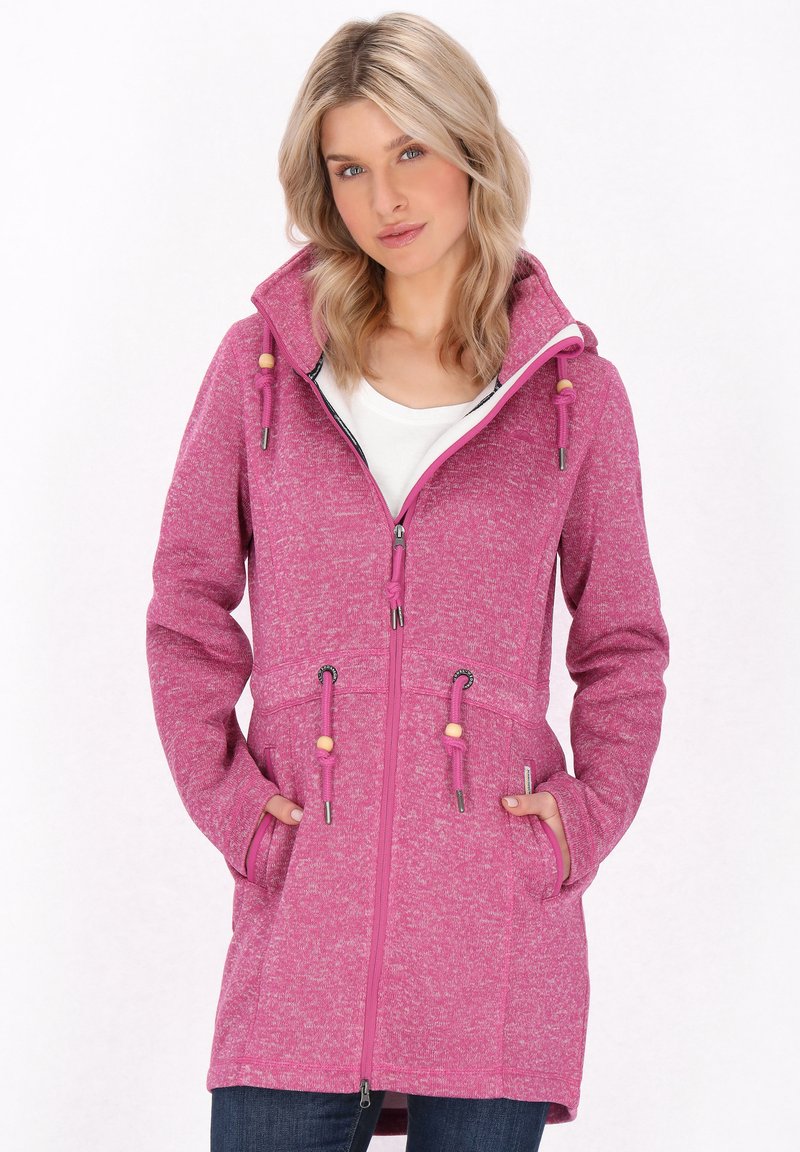 Sieviete, kas valkā garu rozā rāvējslēdzēja kapuci ar auklām un kabatām, apvienojot to ar baltu krekli un tumšiem džinsiem.