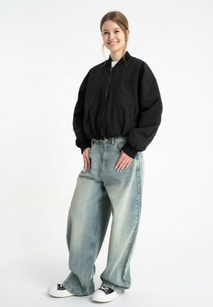 Młoda kobieta uśmiecha się, ubrana w czarną kurtkę na zamek, oversize jasnoniebieskie jeansy i czarno-białe sneakersy na białym tle.
