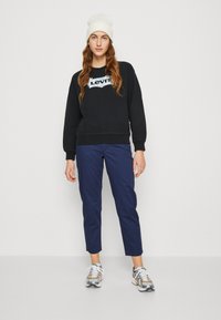 Felpa nera con logo bianco, abbinata a pantaloni blu navy e sneaker argento. Il modello indossa un berretto bianco. Outfit casual, design minimalista.