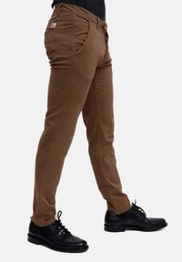 Pantaloni in cotone marrone con vestibilità slim, dotati di tasche laterali e di un'etichetta di branding discreta. Abbinati a scarpe eleganti in pelle nera.