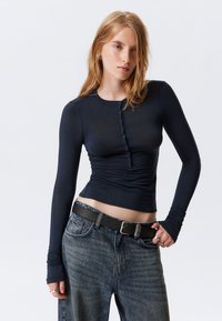Navy long-sleeve top with a high crew neck, featuring a button-down front, ruched sides, and fitted design, paired with high-waisted denim jeans.