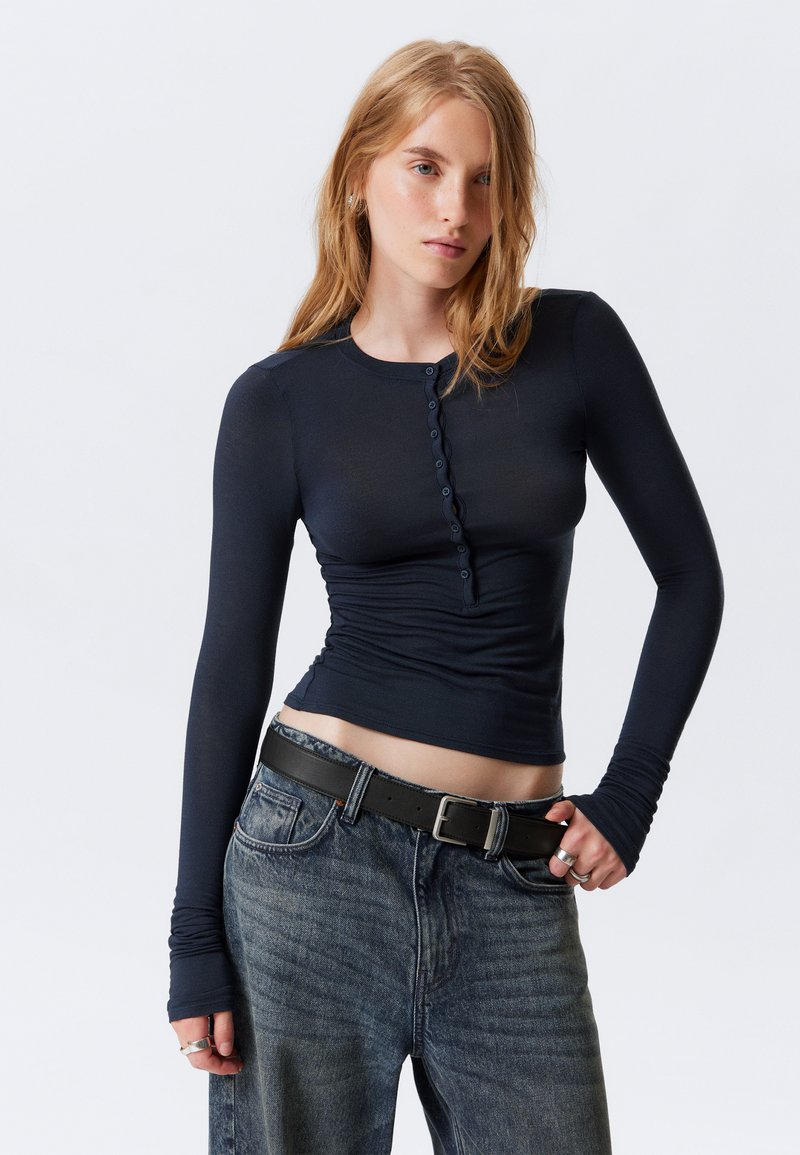 Navy long-sleeve top with a high crew neck, featuring a button-down front, ruched sides, and fitted design, paired with high-waisted denim jeans.
