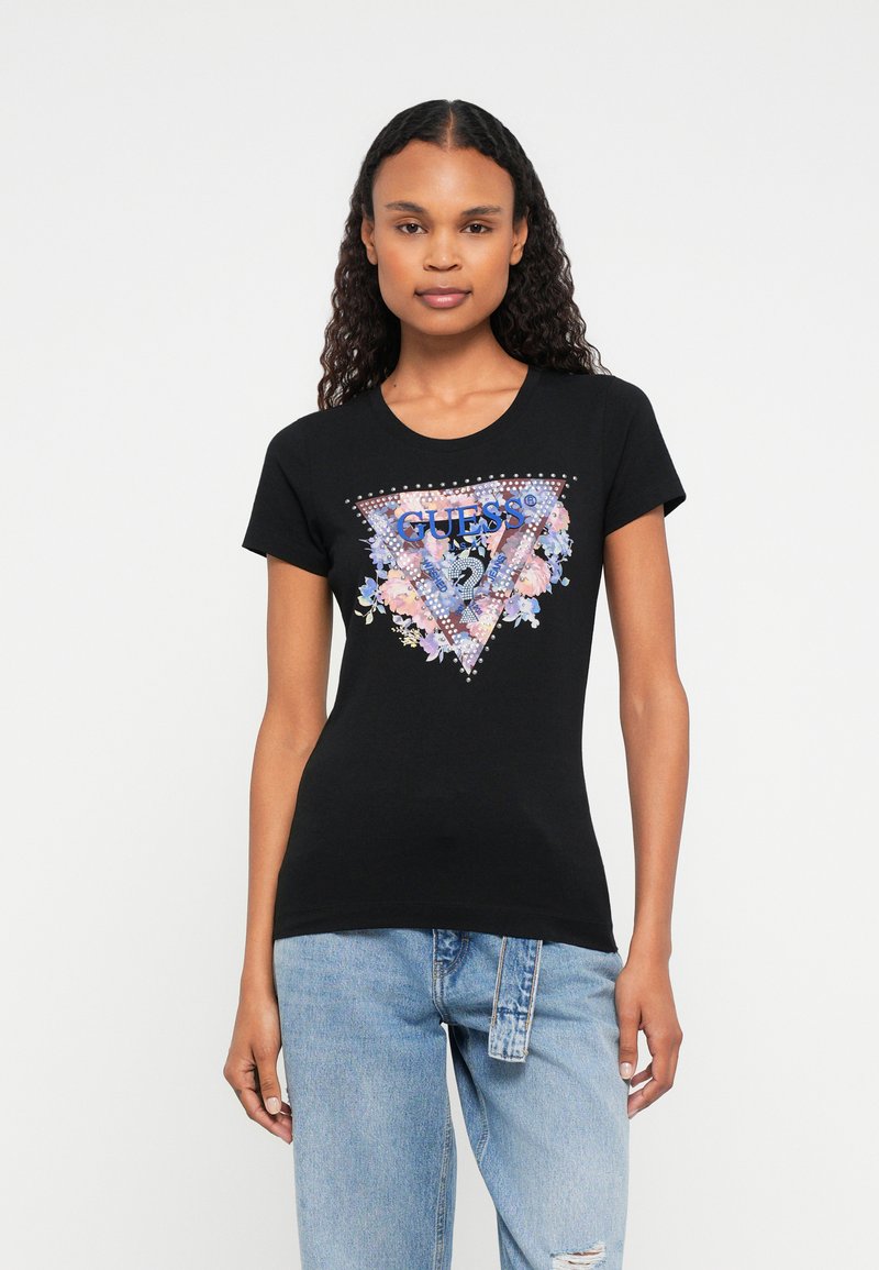 Kvinne som har på seg en svart tettsittende T-skjorte med kort erme med blomstrete Guess-logo og lyseblå høytlivsjeans med stoffbelte.