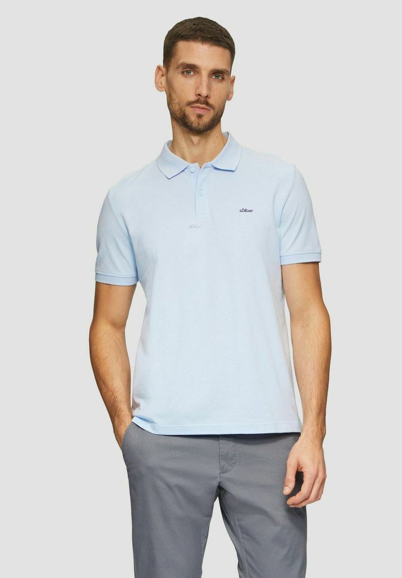 Helles blaues Polohemd aus Baumwolle mit Kragen, kurzen Ärmeln und einem kleinen Logo auf der Brust. Kombiniert mit grauen Hosen.