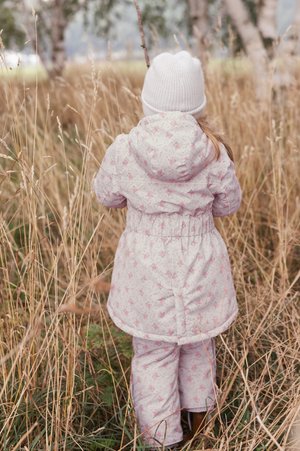 Cappotto imbottito rosa con motivo floreale e cappuccio, abbinato a pantaloni rosa, indossato da un bambino in piedi tra l'erba alta.