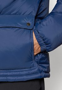 Navy blue puffer jacket featuring a snap closure, elastic cuffs, and side pockets. Smooth, shiny texture with visible stitching details.