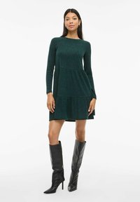 Robe verte à côtes avec manches longues et design à étages. Associée à des bottes noires ajustées jusqu'aux genoux. Encolure ronde simple.