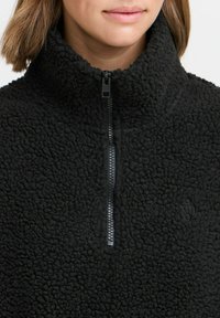 Black fleece pullover with high collar, zip closure, and textured fabric. Features a small logo on the front. Soft, warm appearance.