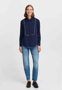 Camicia a maniche lunghe blu navy con cuciture bianche a contrasto, collo e chiusura con bottoni, abbinata a jeans azzurri e mocassini neri.