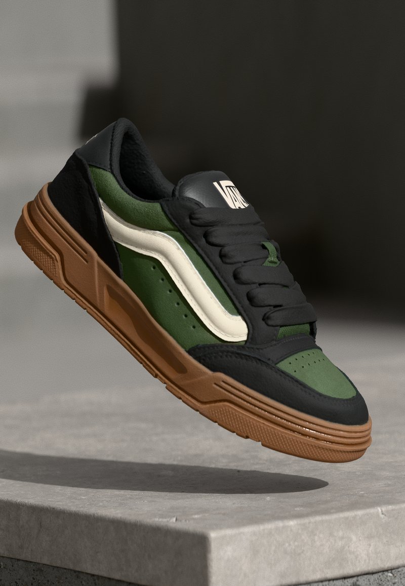 Sneaker med overdel i sort læder, grøn ruskind, hvid bølgelogo og brun gummisål. Har rund tå og snørrestrik.