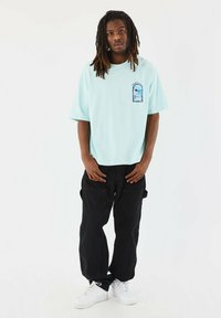 Camiseta oversized de color azul claro con un diseño gráfico en el bolsillo frontal, combinada con pantalones cargo negros y zapatillas blancas.