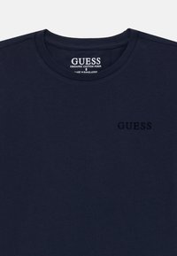 T-shirt en coton bio marine avec col rond, arborant un subtil logo « GUESS » en relief sur la poitrine. Texture douce et design simple.